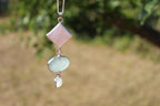 A silver pendant necklace featuring a pink diamond-shaped rose quartz stone, a light green Aventurine oval stone, and a small cushion moonstone, shown against a blurred outdoor background of green foliage and dry grass.